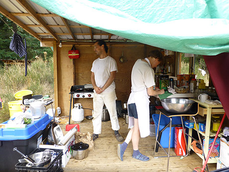 two figures in a makeshift outdoor kitchen, standing amidst camping stoves, rubbermaid tubs and spices