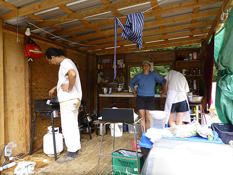 three figures in a makeshift outdoor kitchen, standing amidst camping stoves, rubbermaid tubs and spices