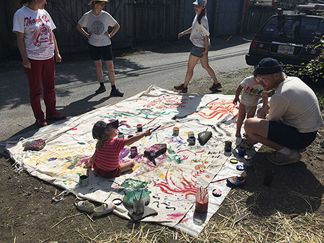 four adults standing around two kids on a large piece of fabric outside in the garden. The canvas has been painted by the kids and adults. there are paints and brushes around the fabric. 