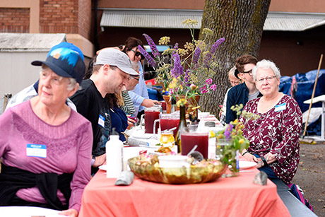 outdoor long table with a dozen adults sitting around it, table is full of food, flowers and pitchers with juice. the crowd is listening to someone talk and not focused on the food