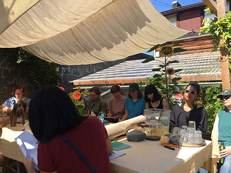 group of people sitting around a long table outside in the garden, with an awning above them for shade. someone seems to be reading and everyone else is following a text. there are dahlias on the table, and two tall sunflowers at the back. the table is full of rocks, flower arrangements, paper, pen, scissors, glassware and lemonade in a rectangular pitcher