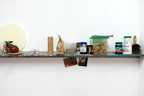 portion of the wall length shelf holding cutting board, pomegrante, photographs, coffee grounds, cookies, pistachios, pumpkin seeds, grape molasses, rosemary cookies, tahini and a brush