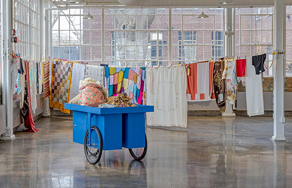 art installation with three laundry lines connecting between poles with two dozen colourful and mutli textured textiles hanging from the lines. sculpture of a tripedal cart with dried flowers and cotton and wool piled on top. the setting is in a large industrial space with big windows and concrete floors