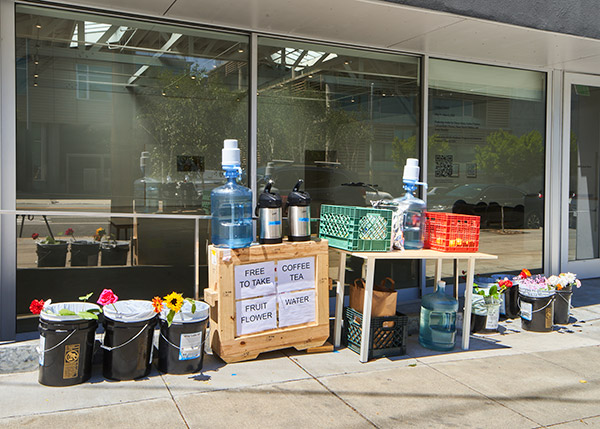 outdoor installation with seven 20 L buckets full of flowers, a wooden crate about 4 feet hight with drinking water and water pump, hot coffee and tea in dispensers, a table with more water and water pumps and two fruit crates full of fruit. there are signs next to the installation saying -Free to Take-, -Coffee and Tea- -Fruit and Flower- and -Water-