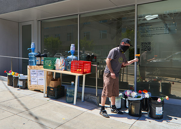 outdoor installation with seven 20 L buckets full of flowers, a wooden crate about 4 feet hight with drinking water and water pump, hot coffee and tea in dispensers, a table with more water and water pumps and two fruit crates full of fruit. there is a person in sneakers shorts and tshirt, black cap and black bandana around their face picking a flower from one of the buckets