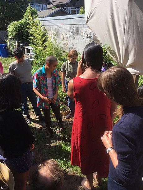 group of people circled around a figure in the center, who seems to be looking at something on the ground and talking. everyone around is listening and looking at the same things. everyone is outside in the garden.