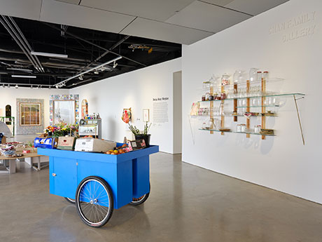 Exhibition view of Meydan, blue cart in the forground and a floating wood and glass shelf on the wall with glassware on it 