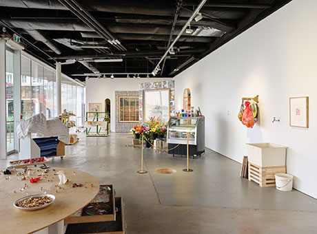 Exhibition view of Meydan with a wooden table, sink in the forground with fridge and cart with blankets in the background