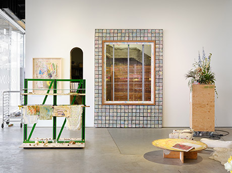 Installation view with window facade sculpture standing in front of sections of the wall cut out making the storage room and the studs of the wall visible