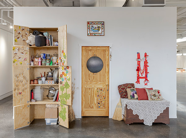 art installation with a 9 foot tall cabinet with paintings of wheat, cotton and hollyhocks painted with watercolours on the inside flaps. the cabinet is full of cooking and food serving equipment, there are a bundle of dired wheat plants leaning on to it, next to it there is a door with a painting of shahmeran and a saj hanging from it, next to the door there is a wooden chest with pillows and a doily on top. on the wall there is a necklace made of red silk ribbon with cold coins and jewelry and money sown on the necklace